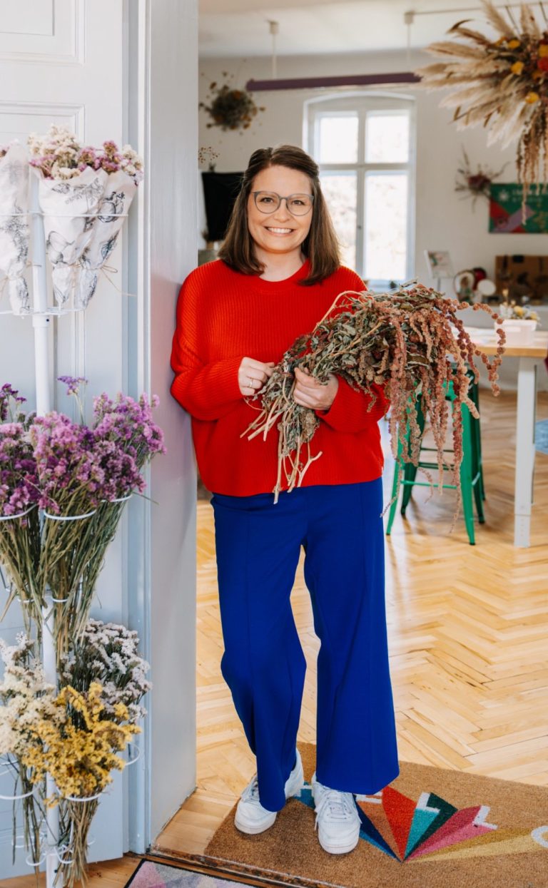 Frau namens Nadin Hoppe mit getrockneten Amaranth auf dem Arm, trägt einen roten Pullover und blaue Hose, lächelt.