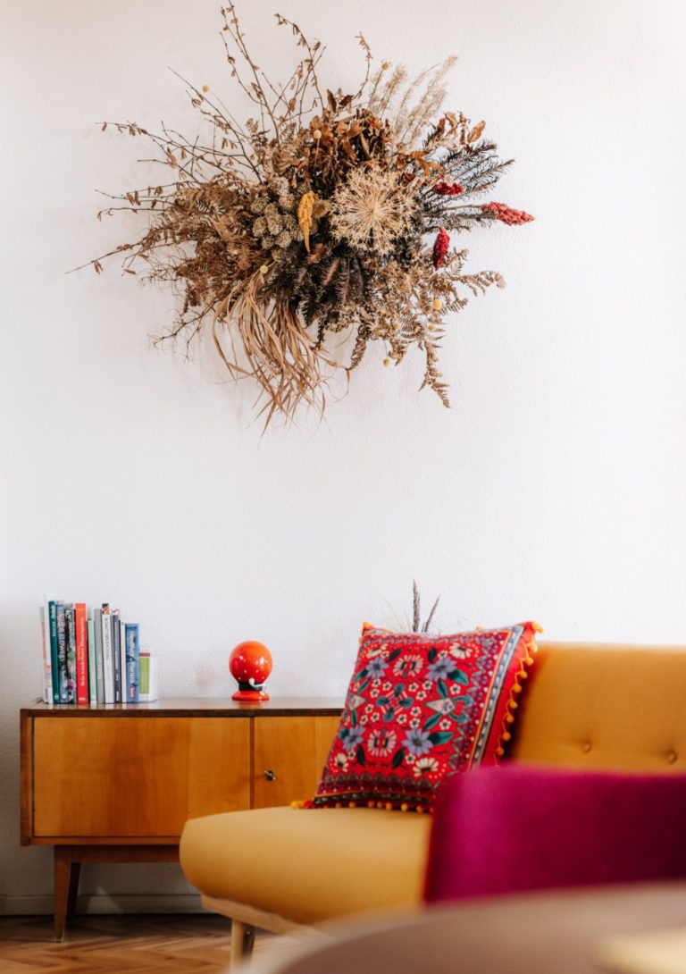 Wanddeko aus Trockenblumen über einem Sofa mit buntem Kissen und Bücherregal.